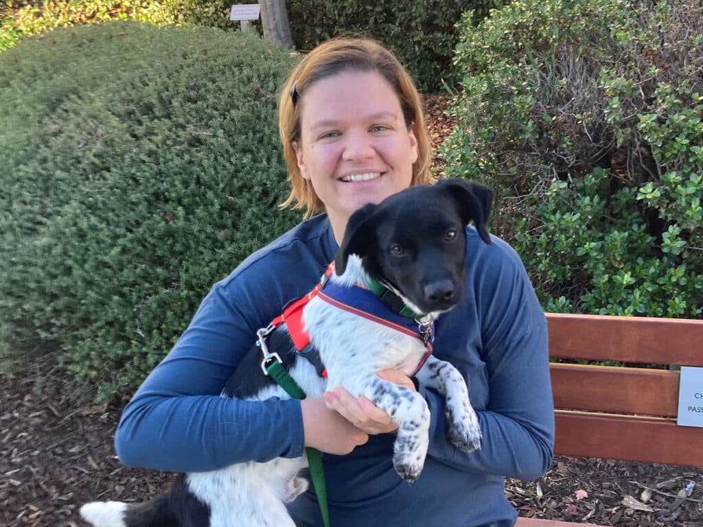 A woman seated on a bench outdoors, smiling while holding a small black and white dog. The dog is wearing a harness, and green foliage is visible in the background, highlighting a joyful moment related to pet adoption.