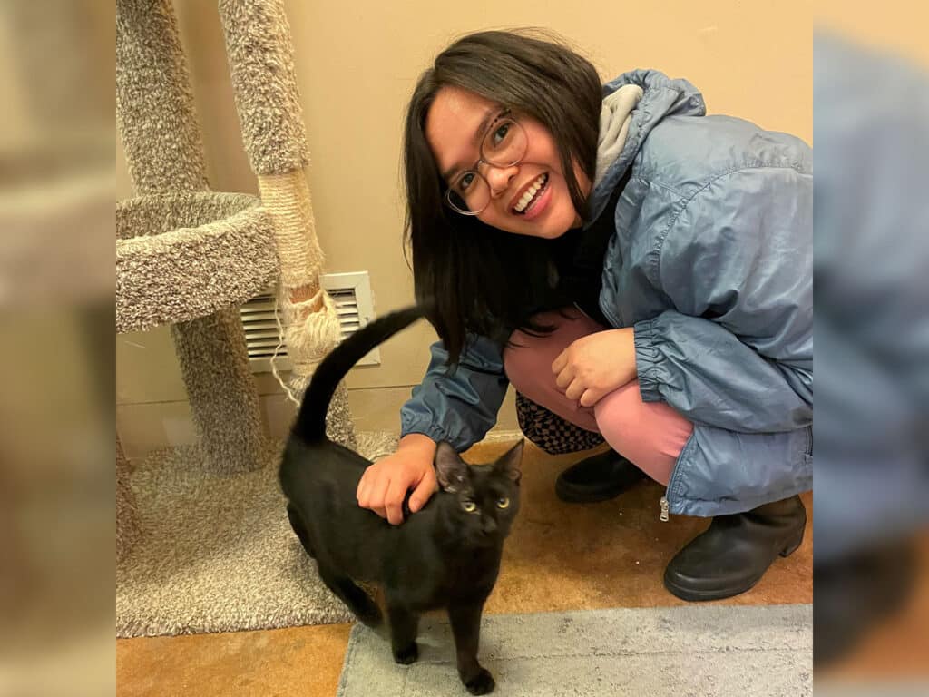 A woman wearing glasses and a blue jacket is crouching beside a black cat. She is smiling and petting the cat, which has its tail raised. The background features a scratching post and a textured floor, creating a cozy setting related to adoption.
