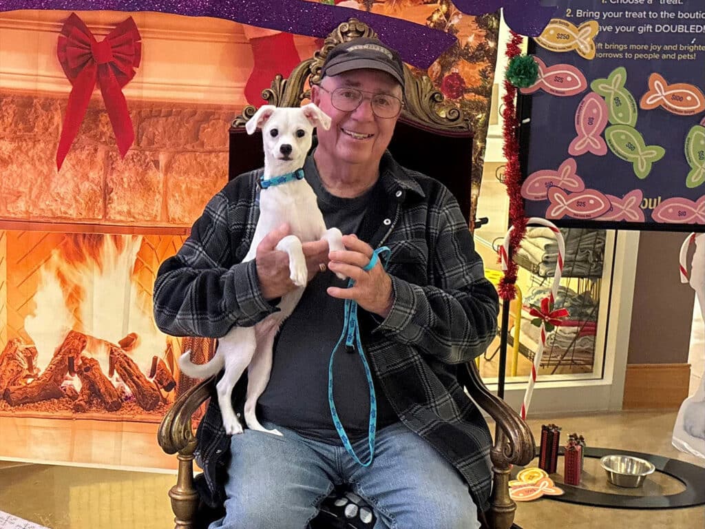 An older man wearing glasses and a cap is seated on a decorative chair, holding a small white dog with a blue collar. The background features a festive setting with a fireplace and holiday decorations. This image relates to the theme of Adoption.