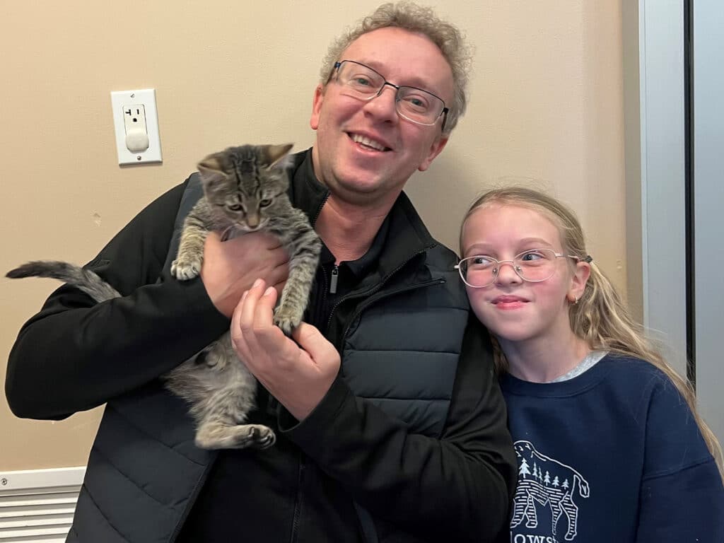 A man and a girl pose together, smiling while holding a gray tabby kitten. The man, wearing glasses and a black vest, stands to the left, while the girl, with glasses and a blue sweatshirt, stands to the right. The background hints at an adoption setting.