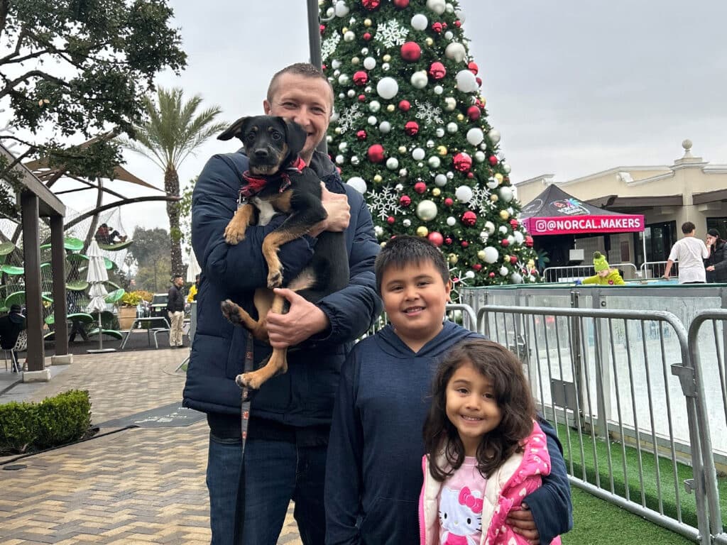 A man holding a small dog stands with two children outdoors, smiling in front of a large decorated Christmas tree. The scene includes palm trees and festive decorations, suggesting a community event related to adoption.