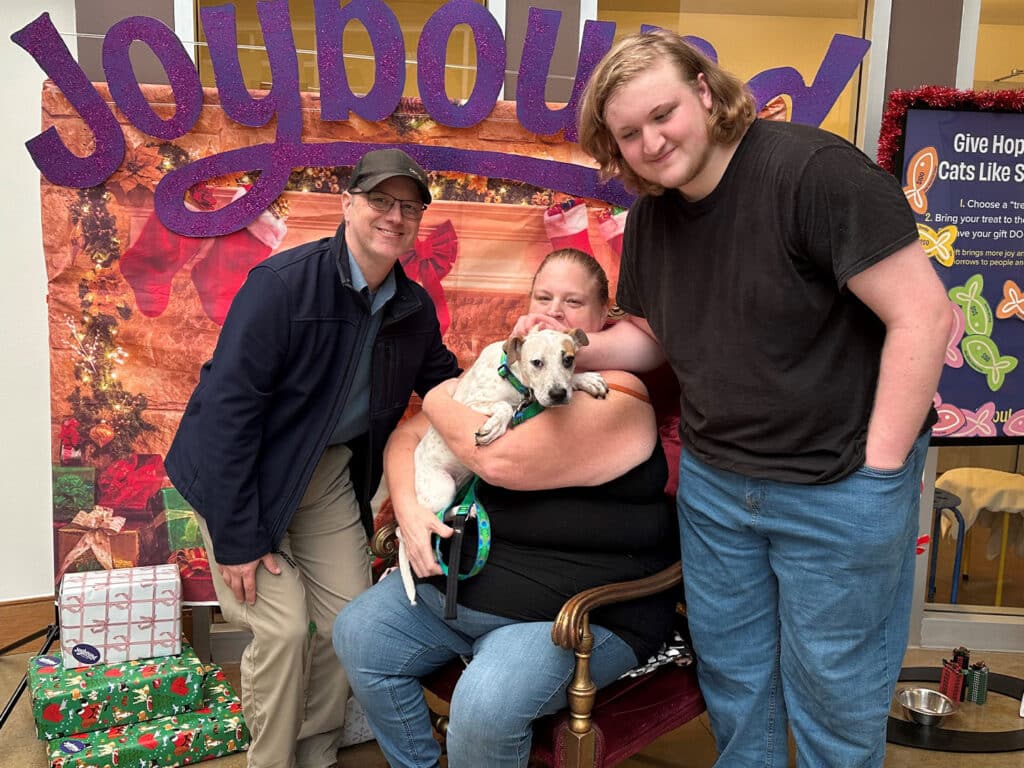 A woman holding a small dog sits on a chair, flanked by a man in a jacket and a young man in a black shirt. They pose in front of a festive backdrop with holiday decorations and wrapped gifts, celebrating pet adoption.