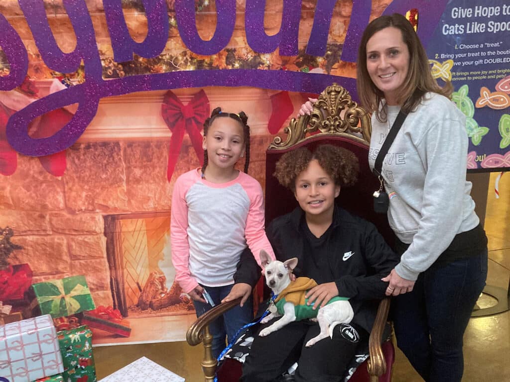 Two children and a woman pose together in front of a festive backdrop. The boy, sitting on a throne, holds a small dog in a green sweater. The girls are smiling, and there are holiday decorations and wrapped gifts in the background, conveying a joyful atmosphere around adoption.