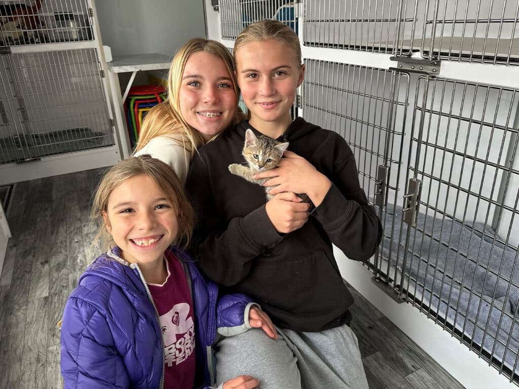 Three young girls smiling while holding a kitten in an animal shelter. The girl in the center is cradling the kitten, while the other two girls are sitting close beside her. Metal crates are visible in the background, indicating an adoption setting.