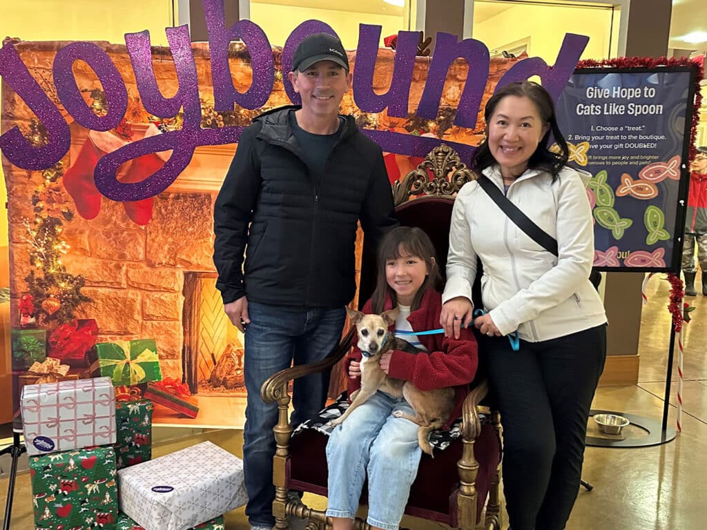 A family poses for a photo during an adoption event, with a man and a woman standing on either side of a girl sitting on a throne-like chair holding a small dog. In the background, a festive decoration reads "Joybound.".