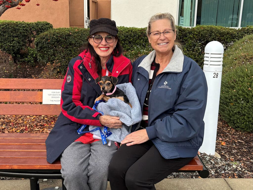 Two women sitting on a wooden bench with a small dog wrapped in a blanket. They are smiling and relaxed, dressed in casual winter attire. A grassy area and building are visible in the background. This image reflects a recent dog adoption.