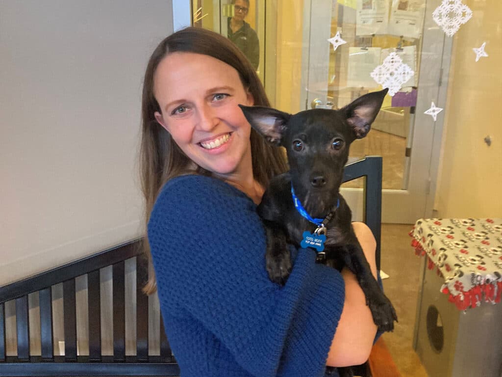 A woman smiles while holding a small black dog with large ears. The setting appears to be a pet adoption center, with soft lighting and visible decorations in the background. The image captures a joyful moment related to pet adoption.