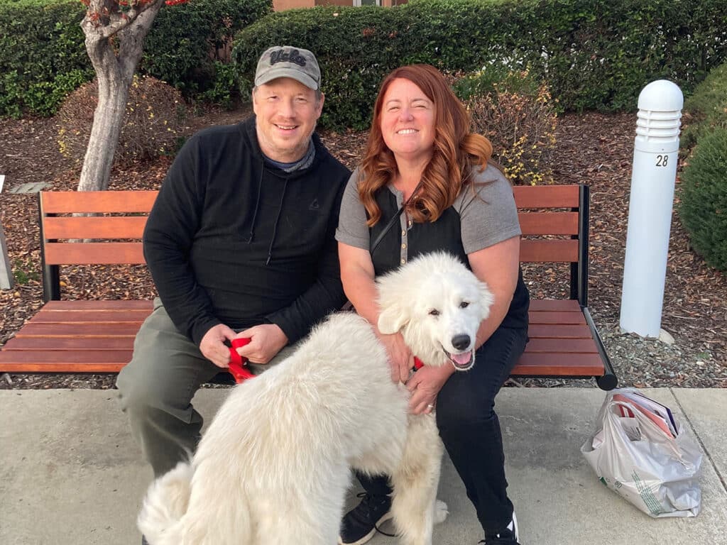 A man and a woman sit on a bench in a park, smiling at the camera. The man has light hair and is wearing a black hoodie and cap. The woman has long, wavy hair and is dressed in a gray shirt. A large, fluffy white dog sits beside them, highlighting their recent adoption.