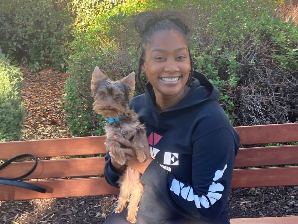A woman sitting on a wooden bench outdoors, holding a small dog in her arms. The dog has a brown and black coat and a blue collar. Surrounding greenery is visible in the background, suggesting a park setting. The image relates to adoption.
