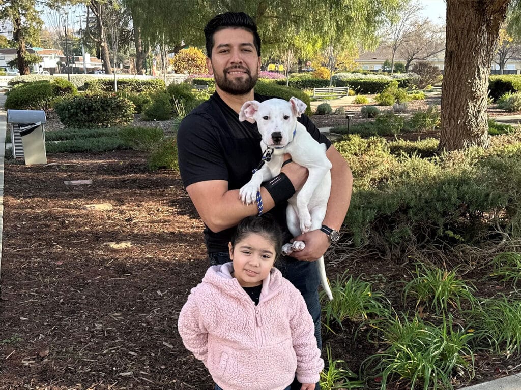 A man holding a small white dog stands in a park with a young girl in a pink jacket beside him. Lush greenery surrounds them, and the background shows trees and park features. This image highlights the joy of pet adoption.