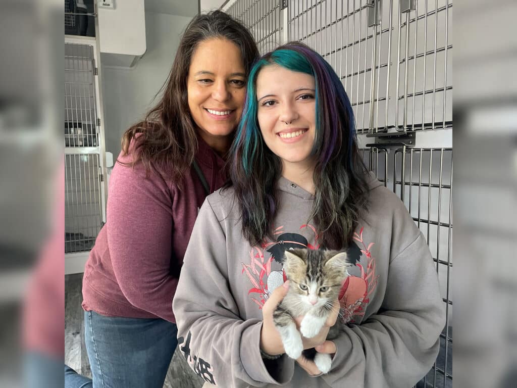 A woman and a young girl pose together in a pet adoption center, smiling. The girl holds a small kitten in her hands. Behind them, there are metal animal cages, indicating a space focused on animal adoption.