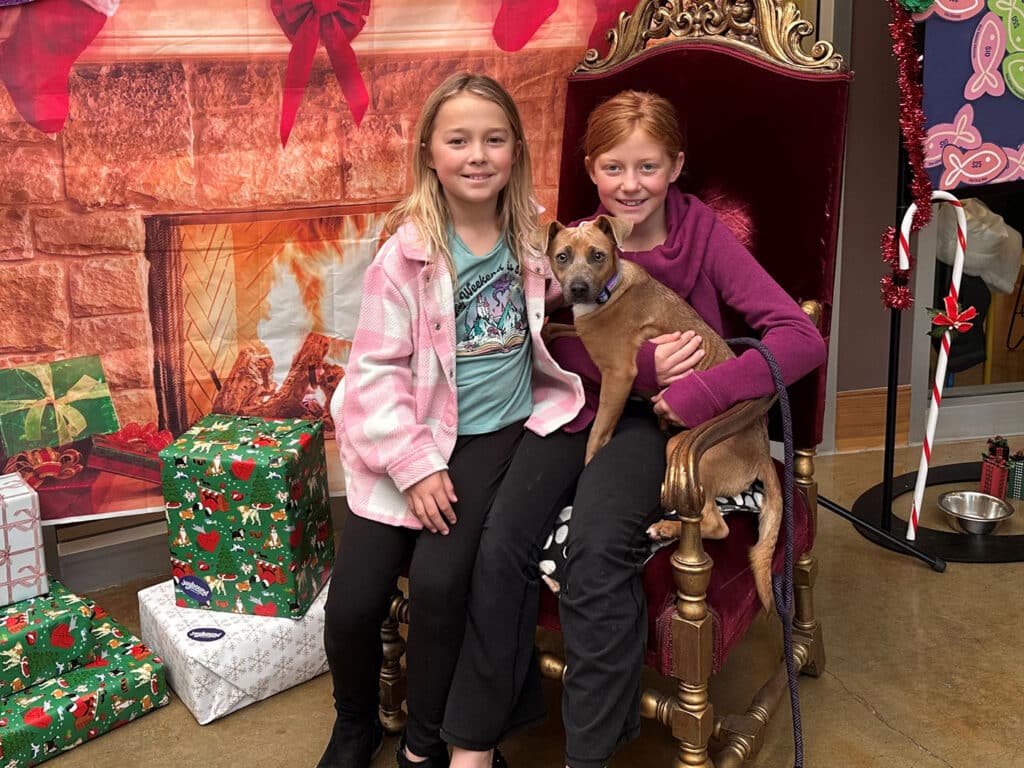 Two girls sitting on a large chair with a festive backdrop, holding a small dog. The scene includes Christmas-themed decorations and wrapped gifts, highlighting a positive moment related to pet adoption.