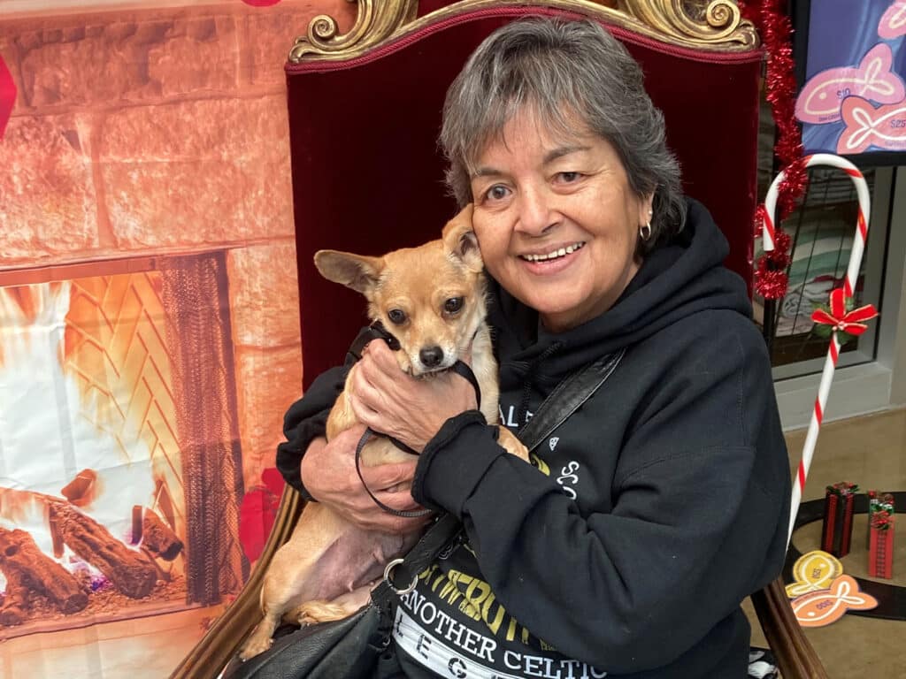 A woman holding a small brown dog on her lap, smiling at the camera. They are positioned in front of a festive background featuring a fireplace and holiday decorations. This image highlights a moment of adoption.