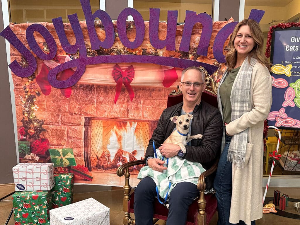 A man holding a small dog sits on a throne-like chair in front of a festive backdrop featuring a fireplace and Christmas decorations. A woman stands beside him, smiling. Wrapped gifts are placed on the floor nearby. The image relates to Adoption.