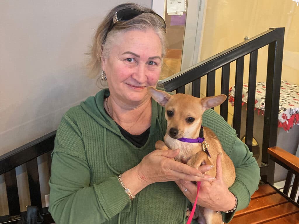 A woman in a green hoodie is holding a small tan dog with large ears. They are seated on a bench, with a wooden background and a visible indoor setting. The scene highlights the joy of adoption.