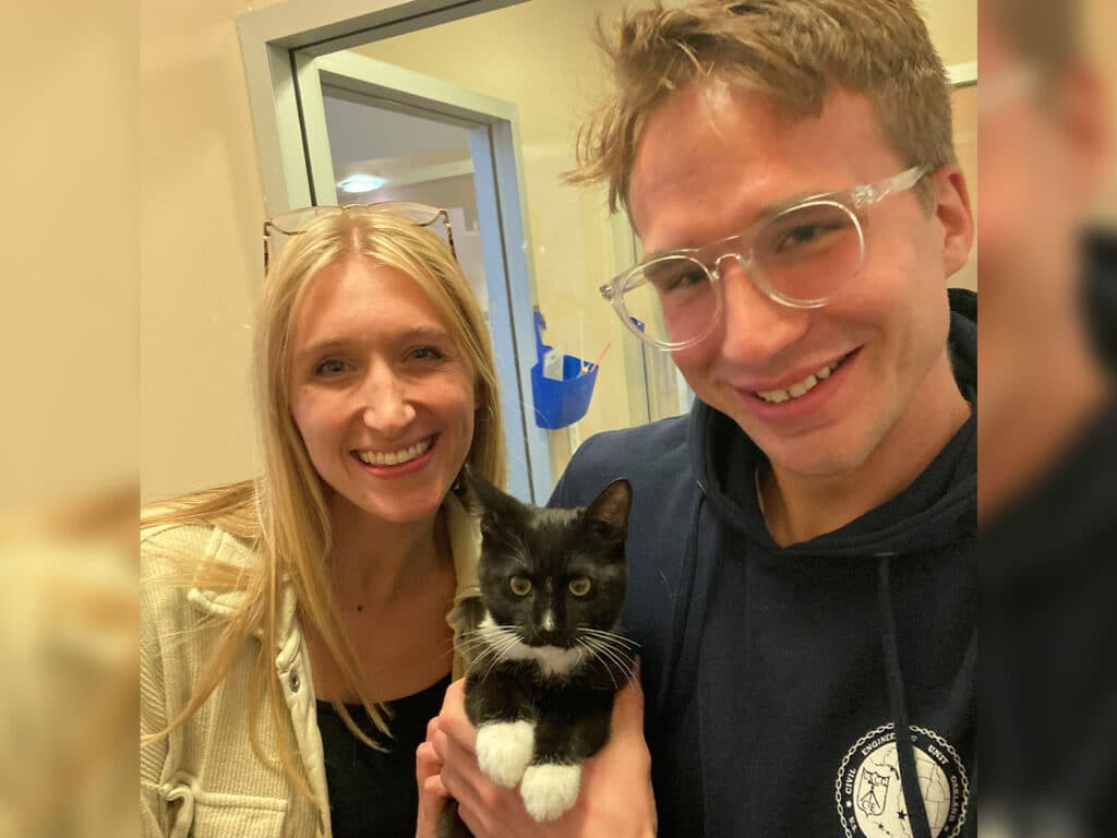 A woman and a man stand together, smiling while holding an adopted kitten with black and white fur. The environment appears to be a pet adoption center, with light-colored walls in the background. This image highlights the joy of adoption.