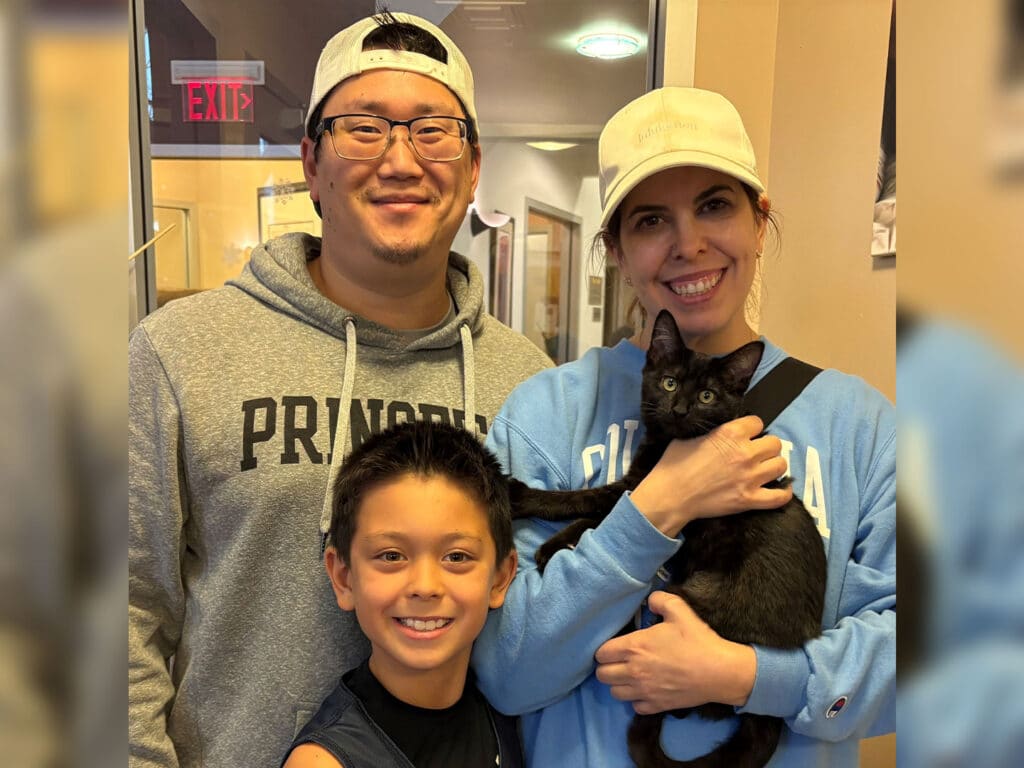 A family of four stands in a shelter, smiling while holding a black cat. The parents, wearing casual sweatshirts and glasses, stand with their son, who is wearing a tank top. The scene reflects a moment of joy following an adoption.