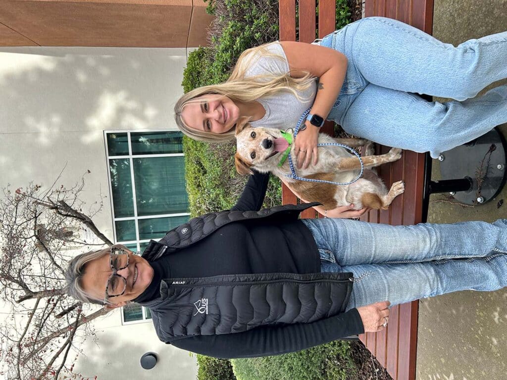 A woman and a young girl seated on a wooden bench outside a building, with a small dog sitting between them. The girl has long hair and is wearing a light shirt and jeans. The woman, dressed in a black vest, smiles and interacts with the dog, representing a recent adoption.