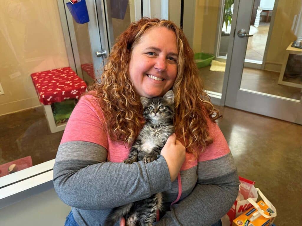 A woman with curly red hair smiles while holding a small gray kitten in her arms. They are inside an animal shelter, with a cozy environment visible, including a red blanket and pet supplies nearby. The image supports the theme of recent adoptions.