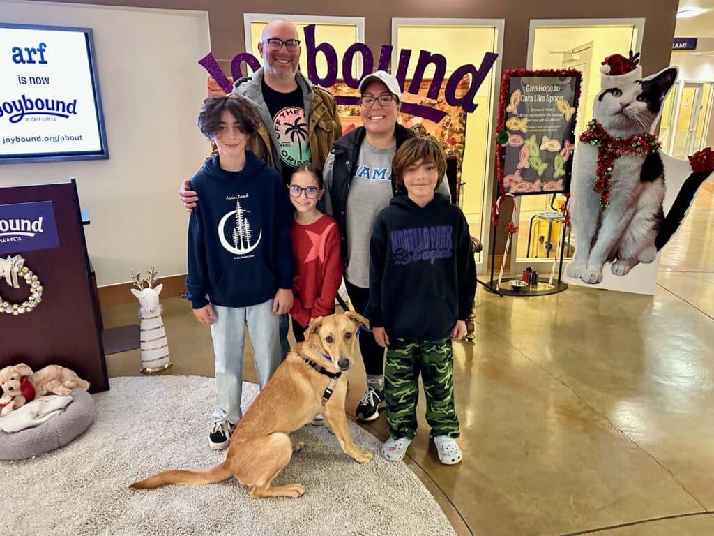 A family of five stands together in a pet adoption center, smiling. A yellow Labrador retriever sits in front of them. The background features signage related to pet adoption, creating a welcoming atmosphere for the event.