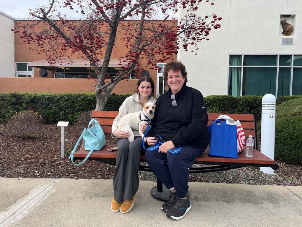 A woman and a man sitting together on a bench, smiling while holding a small dog. The woman has long dark hair and wears a light sweater and gray pants. The background features a tree with red leaves and a building with large windows. Adoption theme.