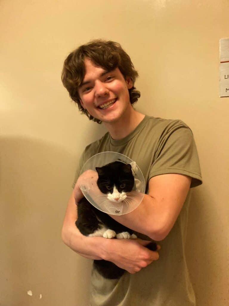 A young man, wearing a gray t-shirt, smiles while holding a black and white cat wearing an Elizabethan collar. The background is a plain wall, and the image highlights the recent adoption of the cat.