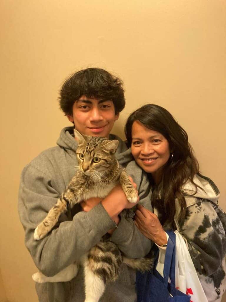 A young man holding a cat in his arms stands next to a woman smiling at the camera. They are in a brightly lit indoor space. Both are dressed casually, and the woman has a bag draped over her shoulder. This image represents a recent adoption.