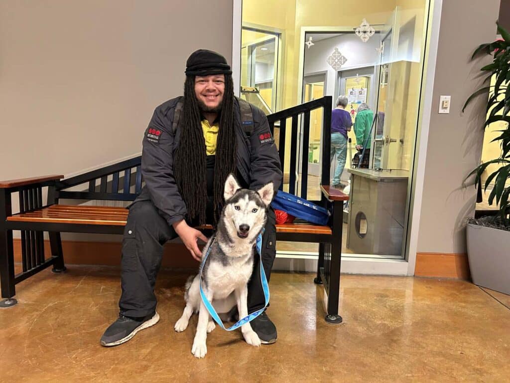 A man with long hair and a black jacket sits next to a gray and white Siberian Husky on a bench. The dog wears a blue leash and appears calm. The background shows an office environment, indicating a recent adoption setting.