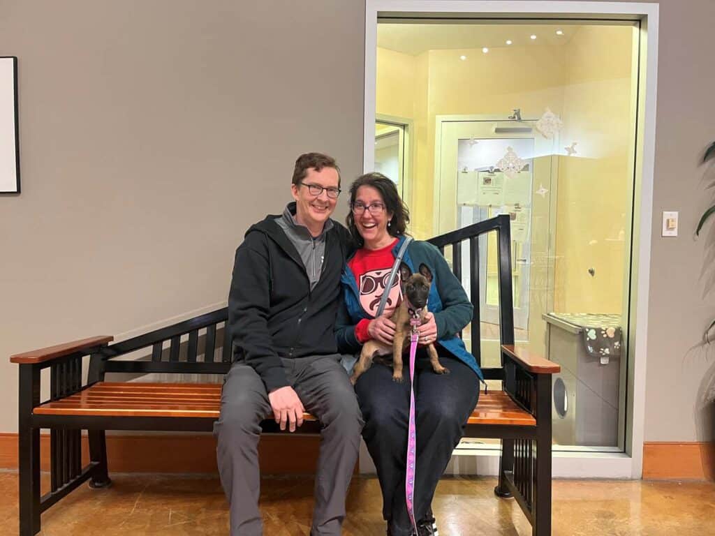 A man and a woman sitting on a wooden bench, smiling while holding a small dog on a pink leash. In the background, a glass door leads to a space with soft lighting. The image highlights the joy of adoption.