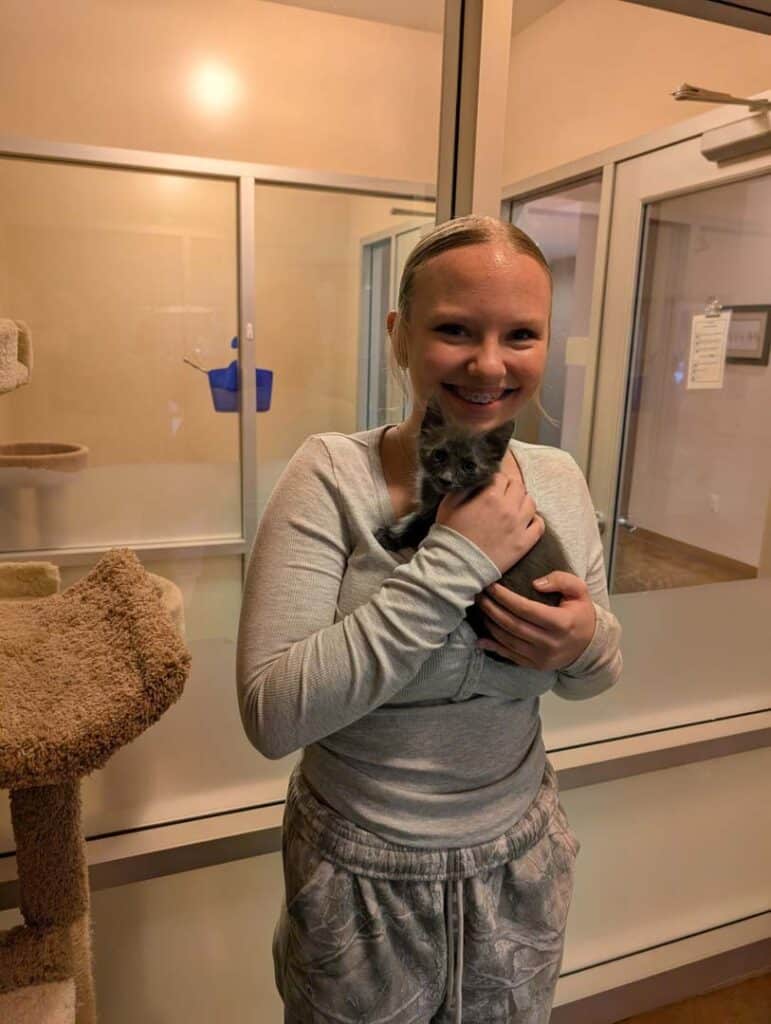 A girl in a light gray outfit holding a small gray kitten in her arms. The background features a cat adoption area with glass panels and a cat tree. This image highlights a moment of joy related to pet adoption.
