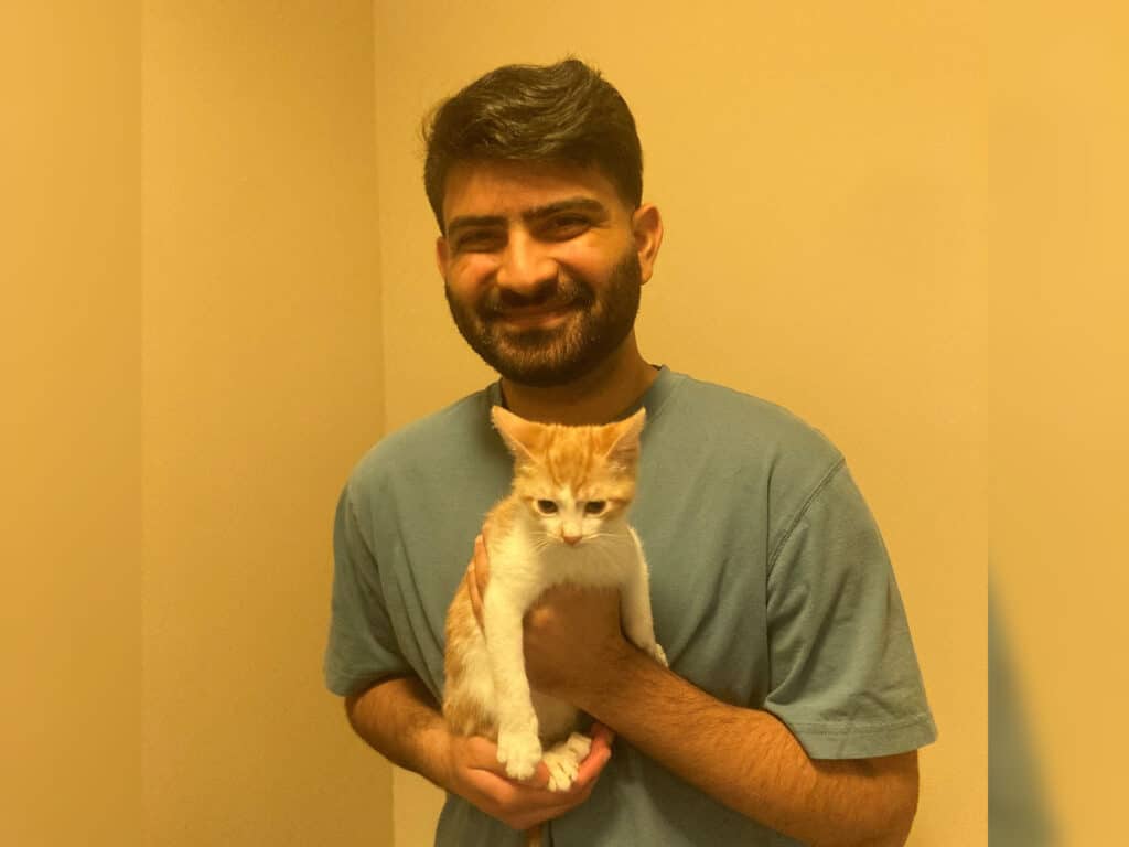 A man with a beard holding an orange and white kitten close to his chest, smiling at the camera. The background is softly lit with neutral colors, suggesting a warm atmosphere. This image highlights a recent adoption experience.