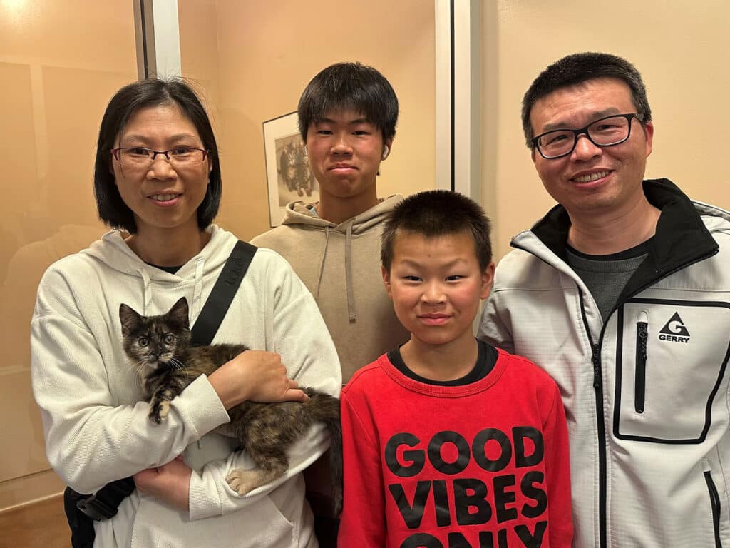 A family of four stands in a brightly lit room. The mother holds a small cat in her arms. Two children, one wearing a red shirt and the other a neutral-colored hoodie, stand beside her. A man in a white jacket, likely the father, stands on the right. Adoption is a central theme.