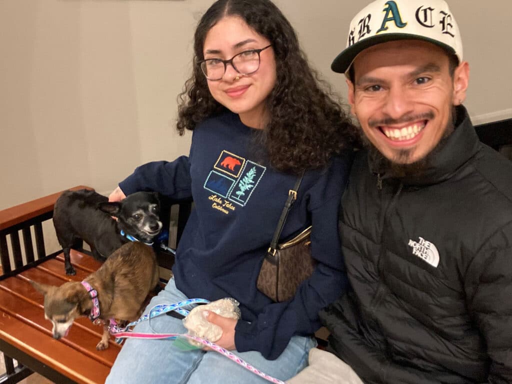 A young woman with glasses and a dark sweater sits on a bench, holding a small dog on her lap. Next to her, a man in a black jacket smiles, also holding a dog on a leash. The scene suggests recent adoptions of these pets.