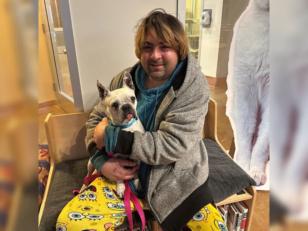 A man with medium-length brown hair, wearing a blue hoodie and gray jacket, sits on a bench, holding a small white dog with black markings. The dog is wearing a pink harness. The man is smiling and wears yellow pants with a cartoon design. This image relates to adoption.
