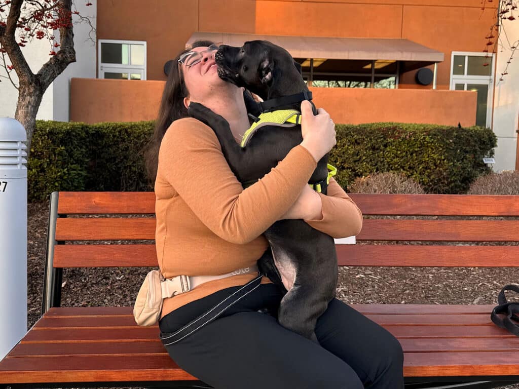 A woman sitting on a wooden bench outside smiles as a black dog with a green harness leans in to give her a kiss. The setting includes shrubs and a building in the background, highlighting a moment of affection likely related to adoption.