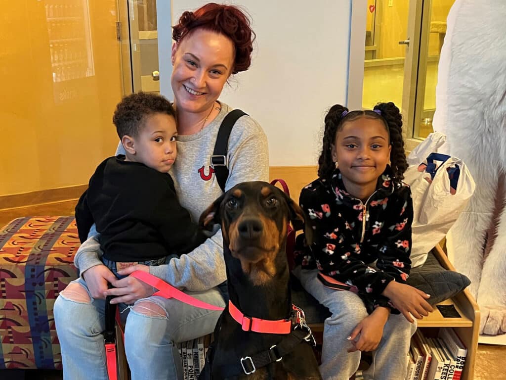 A woman sitting with two children, one on her lap, smiling at the camera. A large Doberman dog is seated in front of them, wearing a red leash. The background features a colorful seating area and large windows, suggesting a welcoming environment for adoption.