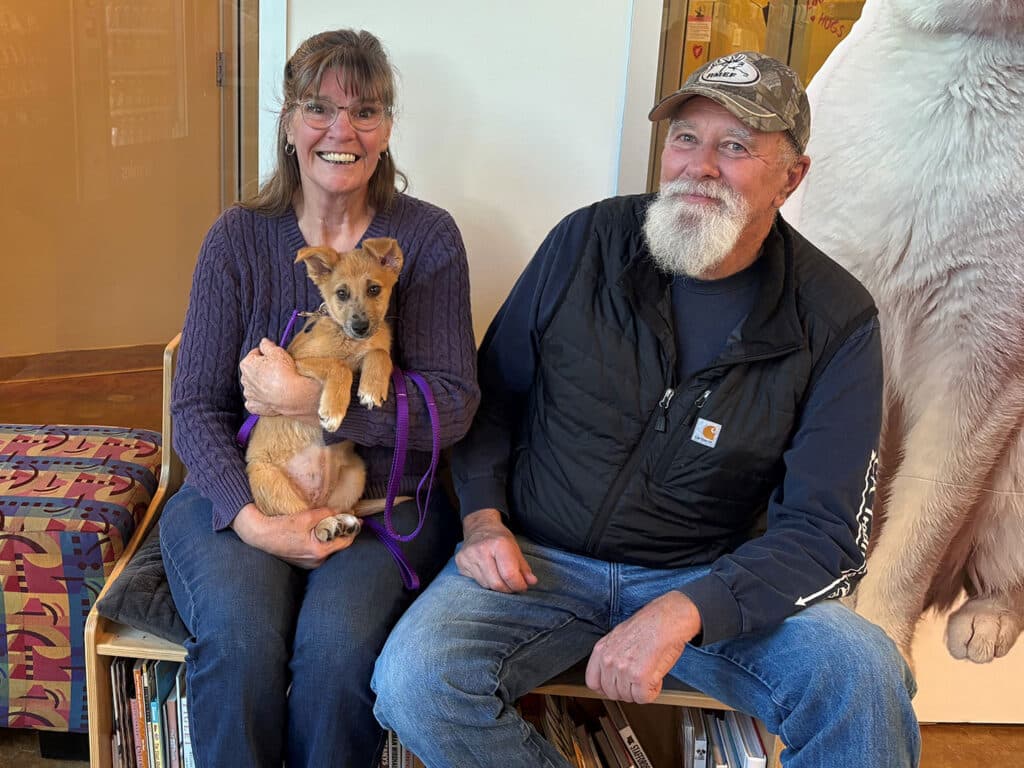 A woman and a man sit together on a bench in a cozy setting, holding a small dog. The woman wears glasses and smiles while holding the dog in her lap, while the man sits beside her, looking at the camera. The image relates to adoption.