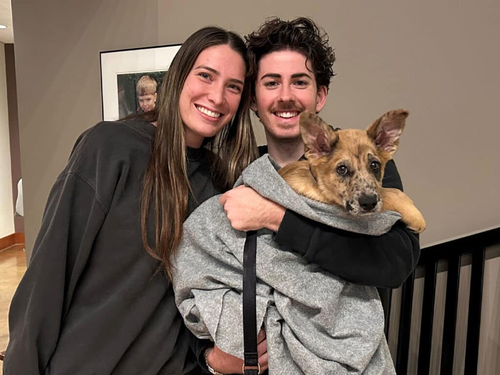 A young couple stands together, smiling while holding a small puppy wrapped in a gray blanket. The setting is indoors with a neutral-colored wall and a framed photograph in the background. The image highlights the joyful moment of pet Adoption.