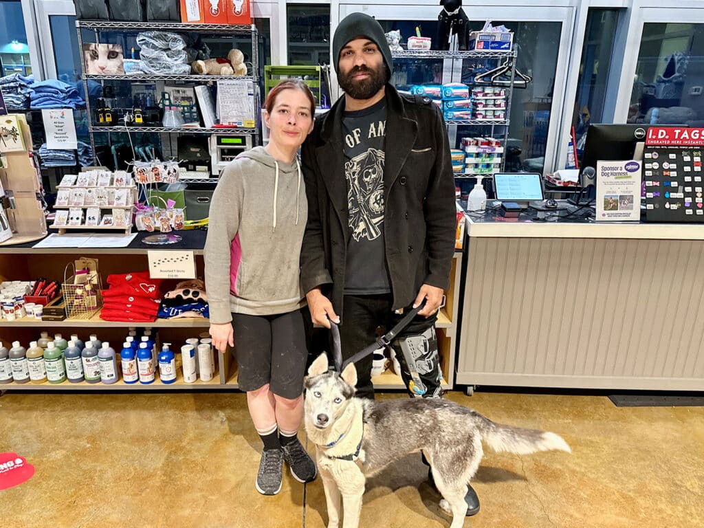 A woman and a man stand together with a dog in a pet store. The woman, wearing a gray hoodie and black shorts, is smiling. The man, dressed in a black coat and beanie, holds the dog’s leash. The background features pet supplies and accessories, highlighting adoption.