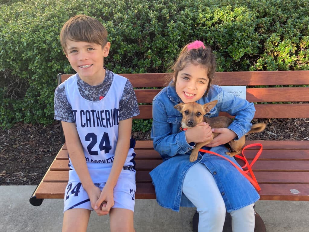 Two children sitting on a bench in a park, a boy wearing a sports jersey on the left and a girl on the right holding a small brown dog. The background features green shrubbery. The image highlights themes of friendship and adoption.