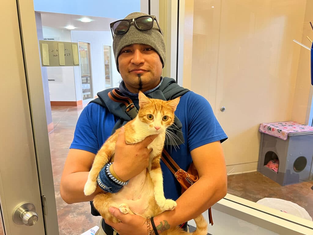 A man wearing a beanie and sunglasses holds an orange and white cat in his arms at an animal shelter. The background shows a clean room with pet facilities. This image highlights a moment of pet adoption.