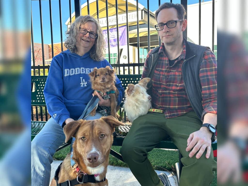 A man and a woman sitting on a bench outdoors with three dogs. The woman wears a blue hoodie and has curly hair, while the man is dressed in a plaid shirt and vest. The dogs, two small breeds and one medium-sized, are seated beside them, showcasing their involvement in the adoption process.