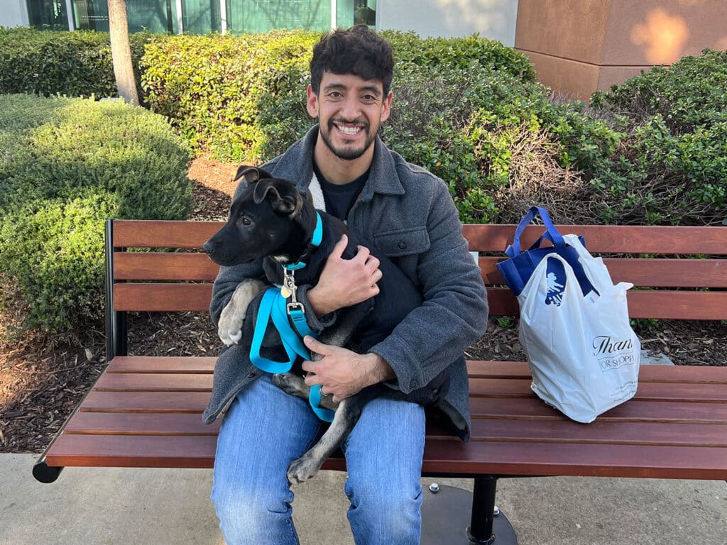 A man sits on a park bench holding a small dog in his arms. The dog is wearing a blue harness and leash. Beside the man, there is a tote bag on the bench. The background features greenery and a building, suggesting a location related to pet adoption.