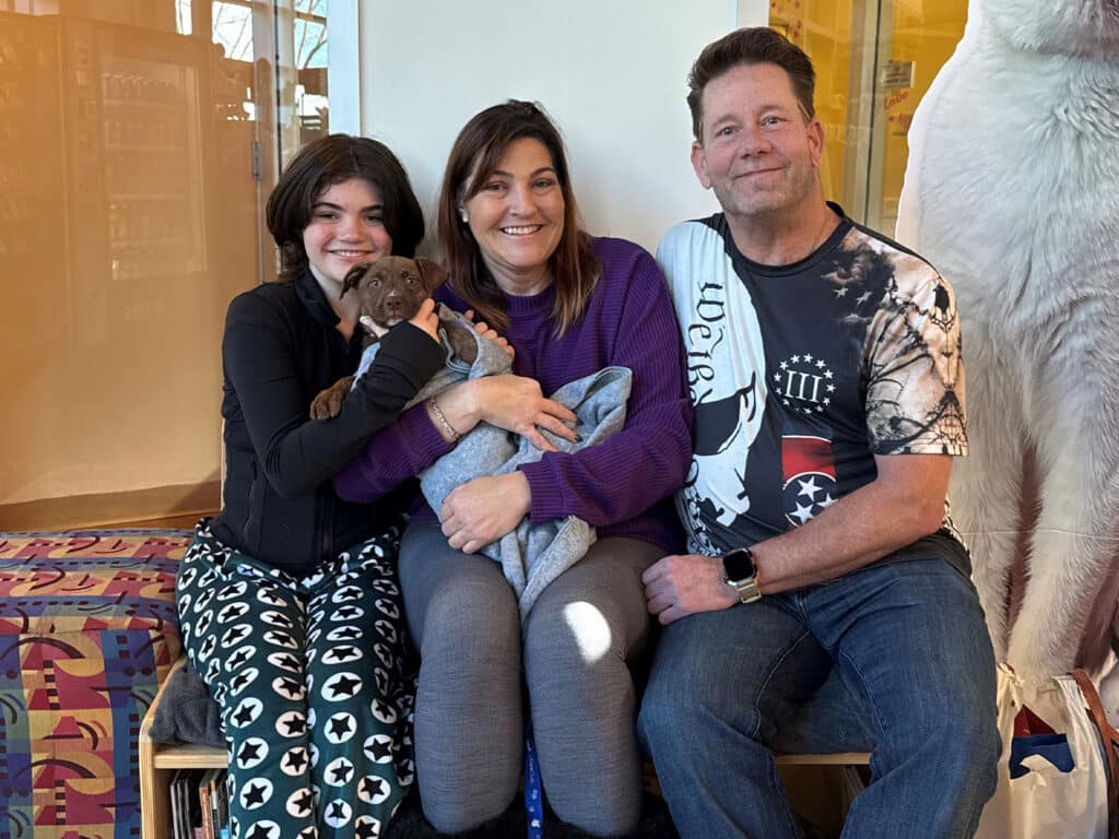 A girl and two adults are sitting together, smiling while holding a small brown puppy wrapped in a blanket. The background shows a colorful, modern space, highlighting the joy of adoption.
