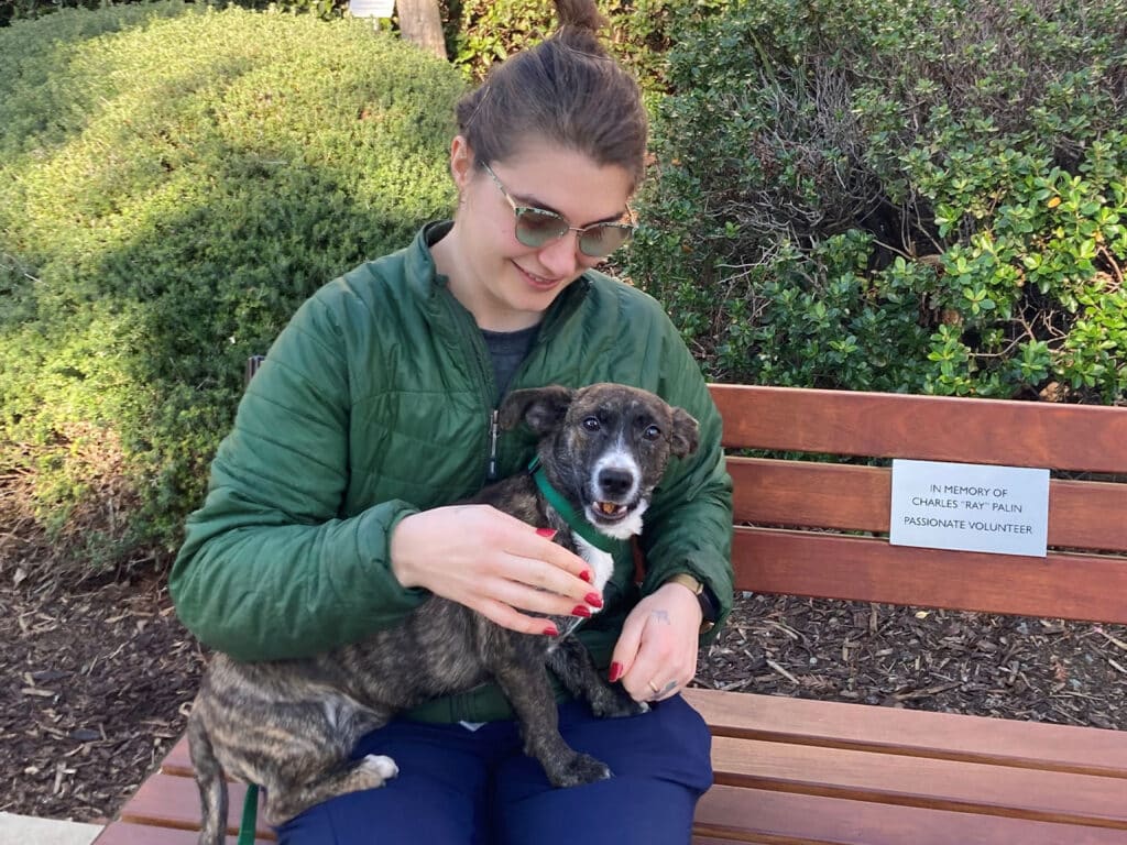 A woman wearing glasses and a green jacket sits on a bench, holding a small brown dog with a playful expression. The background features greenery and a sign honoring volunteers. This image highlights the theme of adoption.
