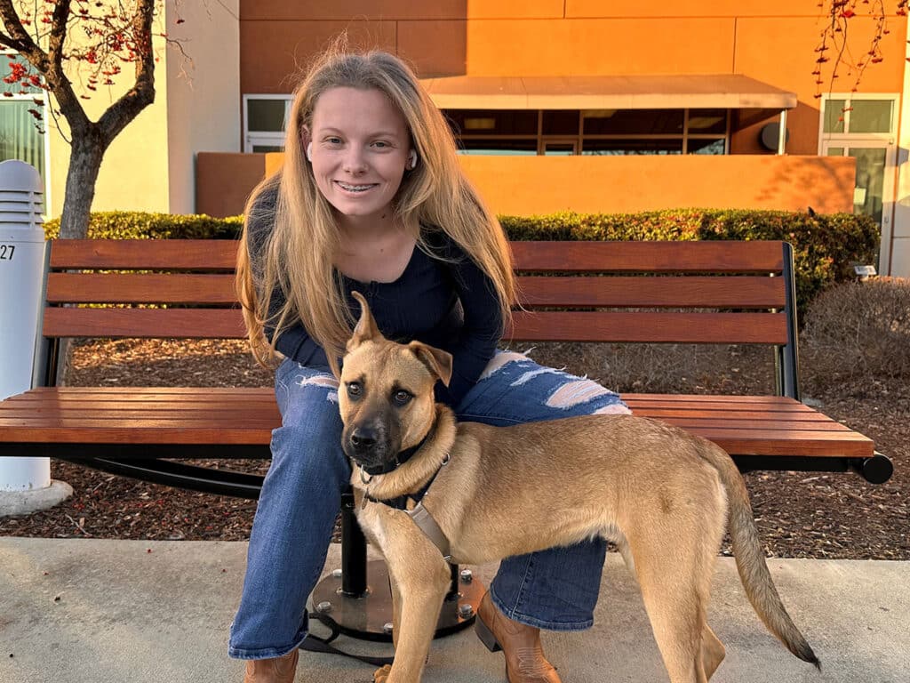 A girl with long blonde hair sits on a park bench beside a light brown dog. The dog looks up at her while she smiles, wearing casual jeans and a black shirt. The background features bushes and a building, highlighting a scene related to pet adoption.