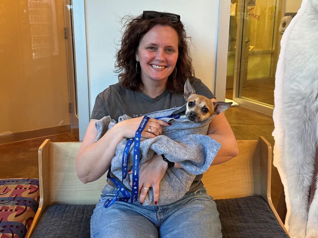 A woman holding a small dog wrapped in a gray blanket while seated on a wooden bench. The woman has dark, curly hair and is wearing a gray t-shirt. The setting appears to be a pet adoption center, suggesting a recent Adoption.