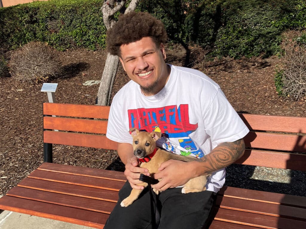 A man sitting on a wooden bench in a park, smiling while holding a small brown puppy with a red collar. The background features trees and landscaped greenery, conveying a joyful moment related to adoption.