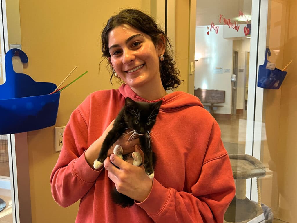 A young woman smiling while holding a black and white kitten in a pet adoption center. The background features a glass door and colorful storage bins. This image highlights the joyful experience associated with pet adoption.