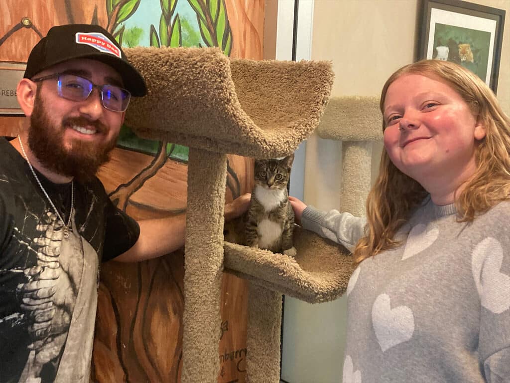 A man and a woman stand beside a cat tree, with a gray tabby cat visible on a middle platform. The woman smiles at the camera while the man leans in close. They are in a bright, welcoming environment, likely a shelter, celebrating the adoption.